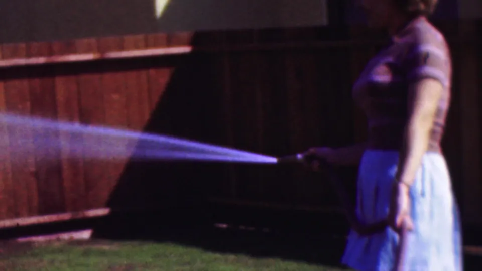 1953 Cleveland 8mm frame showing a woman standing near the right edge of the frame as she waters grass beside a wooden fence.