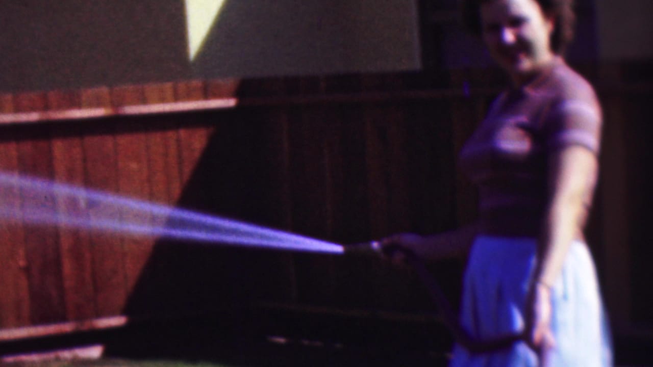In this 1953 Cleveland home-movie frame, a woman in a striped short-sleeve top and light skirt smiles while directing a garden hose across the yard near a wooden fence.