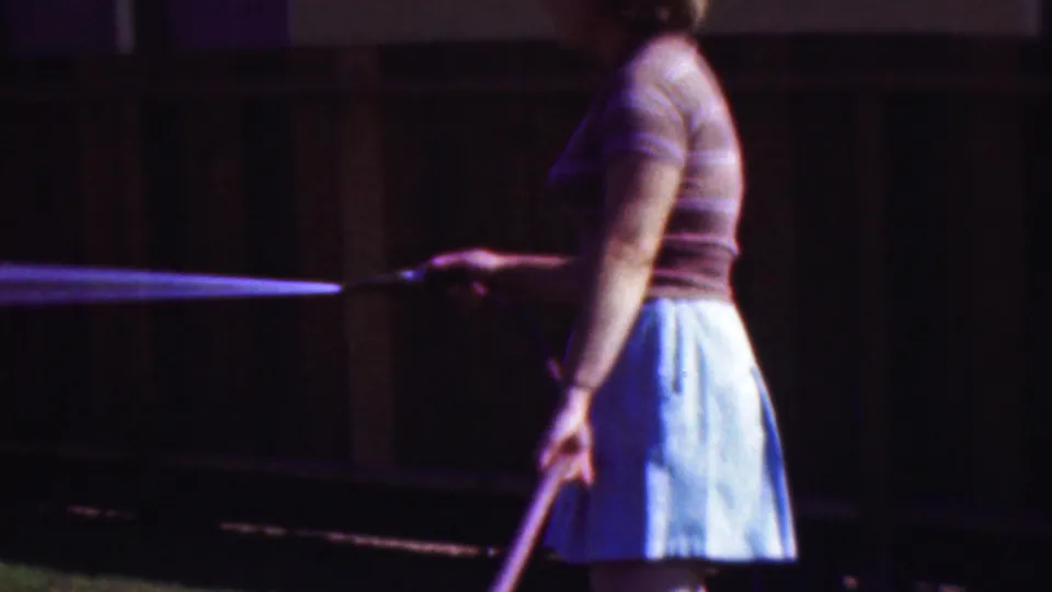 The woman turns sideways in the yard while continuing to water the grass, with the garden hose, water spray, light skirt, and dark wooden fence visible in the soft 8mm frame.