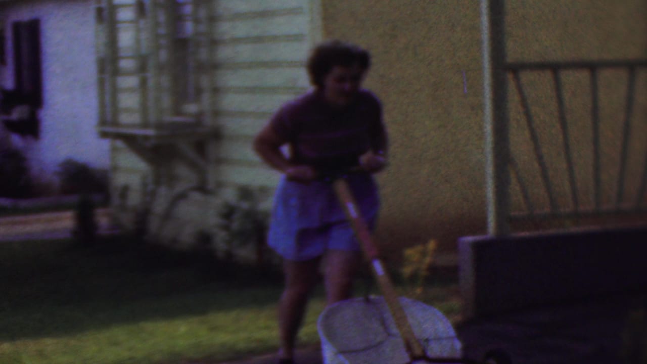 In this 1953 Cleveland home-movie frame, a woman in a striped short-sleeve top and light blue bottoms pushes a wheelbarrow across a grassy yard beside a light-sided house.