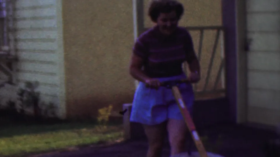 The woman smiles while handling the wheelbarrow in the Cleveland yard, with house siding, a dark doorway or porch area, and lawn visible behind her.