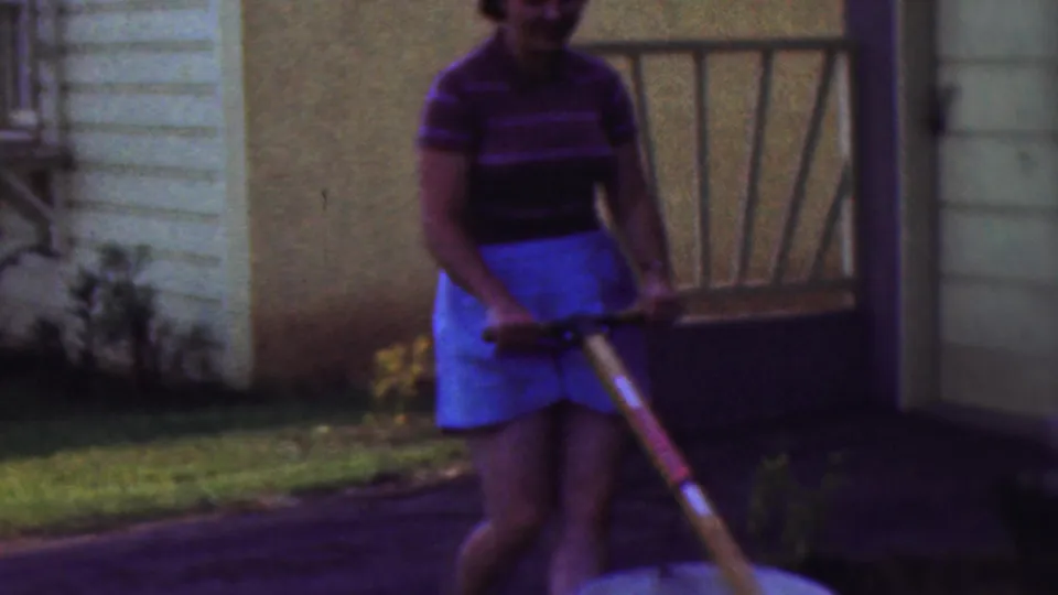 A closer 1953 Cleveland frame shows the woman guiding the wheelbarrow across the lawn, with light-colored siding and a porch or doorway structure in the background.