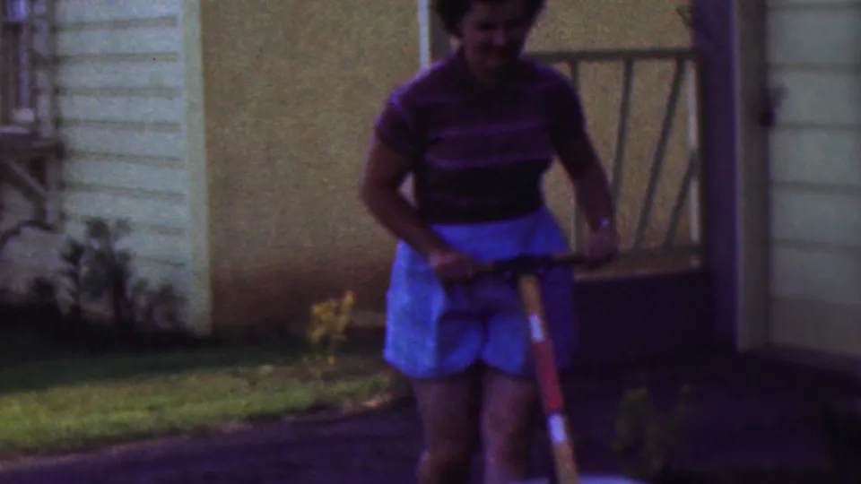 The final frame shows the woman smiling while holding the wheelbarrow handles in the 1953 Cleveland yard, with grass, shrubs, house siding, and a doorway area behind her.