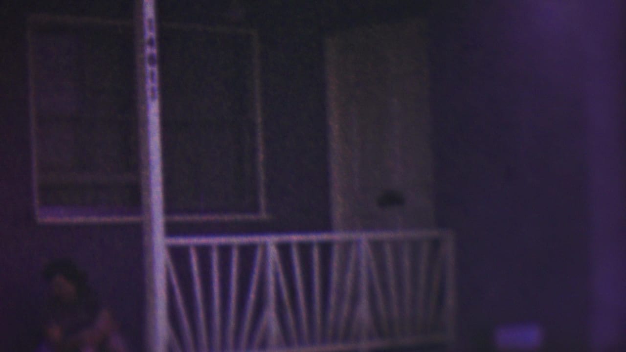 A woman sits in shadow near the front stoop of a Cleveland house in 1953, with a white railing, doorway, and windows visible under purple-blue low-light 8mm color.