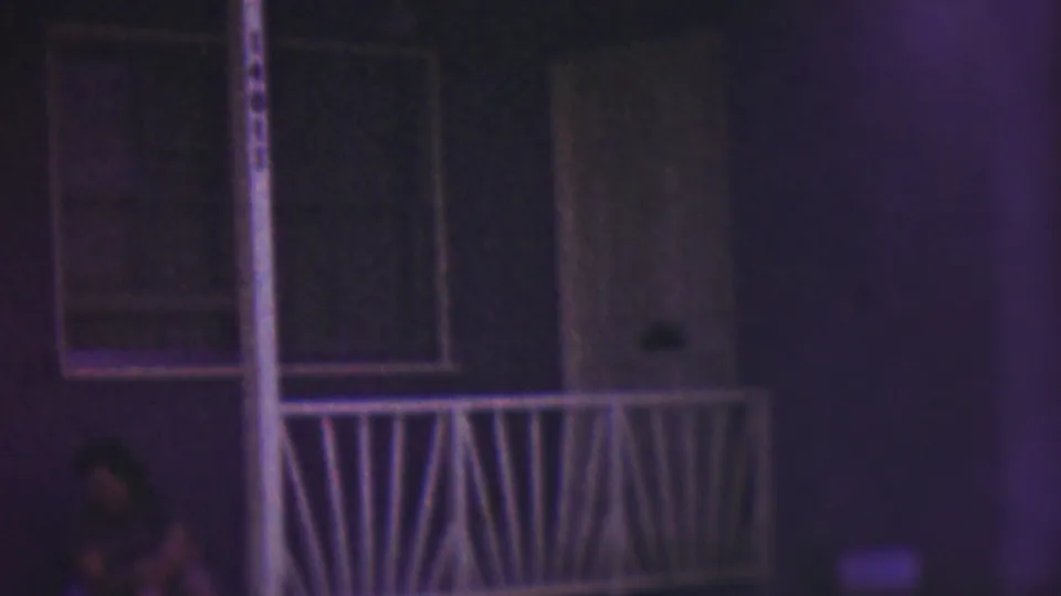 A woman sits in shadow near the front stoop of a Cleveland house in 1953, with a white railing, doorway, and windows visible under purple-blue low-light 8mm color.