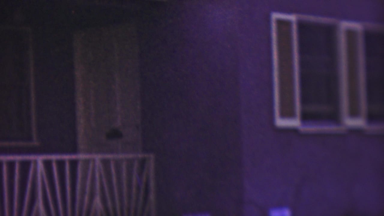 The camera frames the Cleveland residential exterior with a front door, porch railing, and windows, while the seated woman is mostly outside the clear center of the frame.