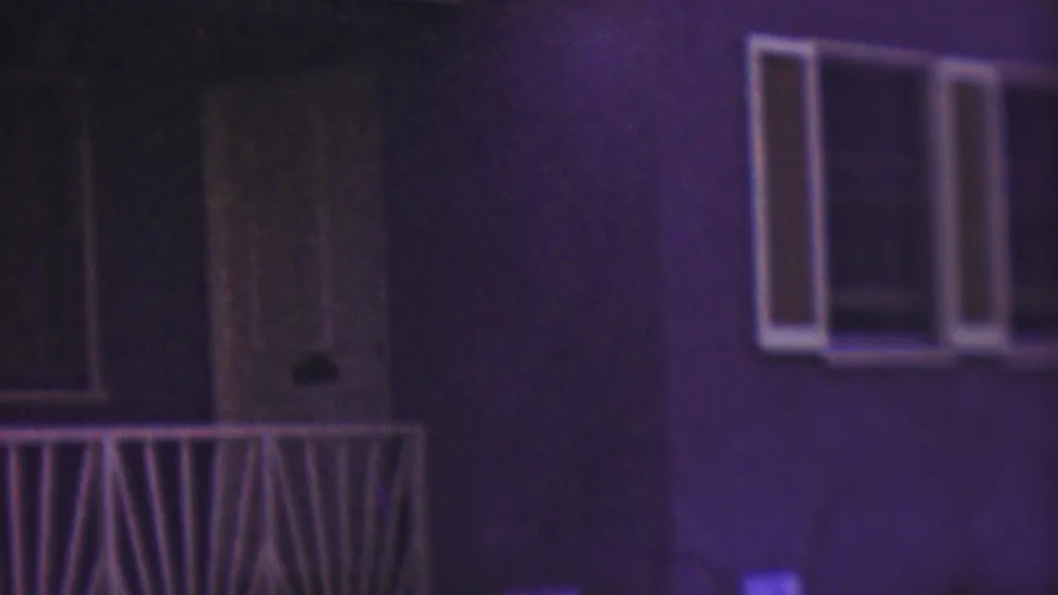 The camera frames the Cleveland residential exterior with a front door, porch railing, and windows, while the seated woman is mostly outside the clear center of the frame.