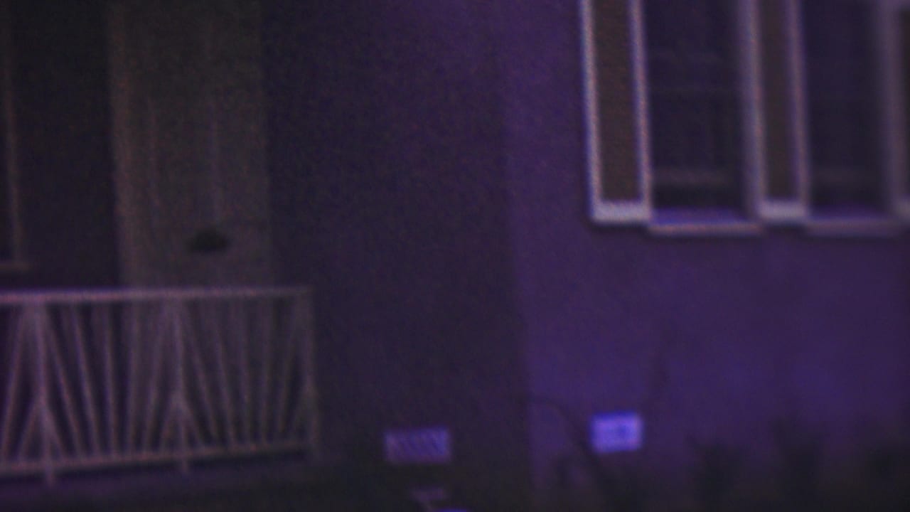A low-light 8mm view of the house facade shows white porch railing, a window, steps, and dark landscaping or yard area, with the figure only faintly visible near the left edge.