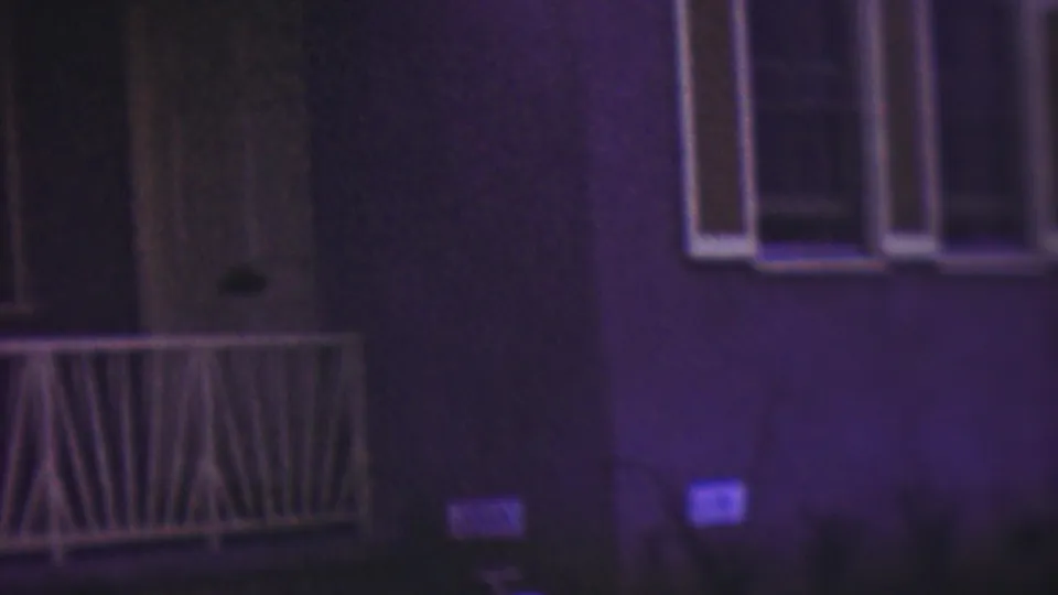 A low-light 8mm view of the house facade shows white porch railing, a window, steps, and dark landscaping or yard area, with the figure only faintly visible near the left edge.
