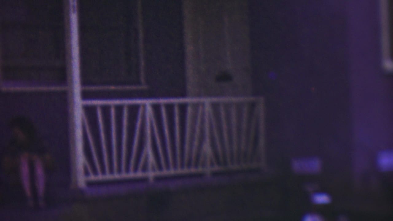 The woman is more centered on the front steps of the Cleveland house, seated beside the porch railing and doorway in soft, grainy 1953 8mm nighttime footage.
