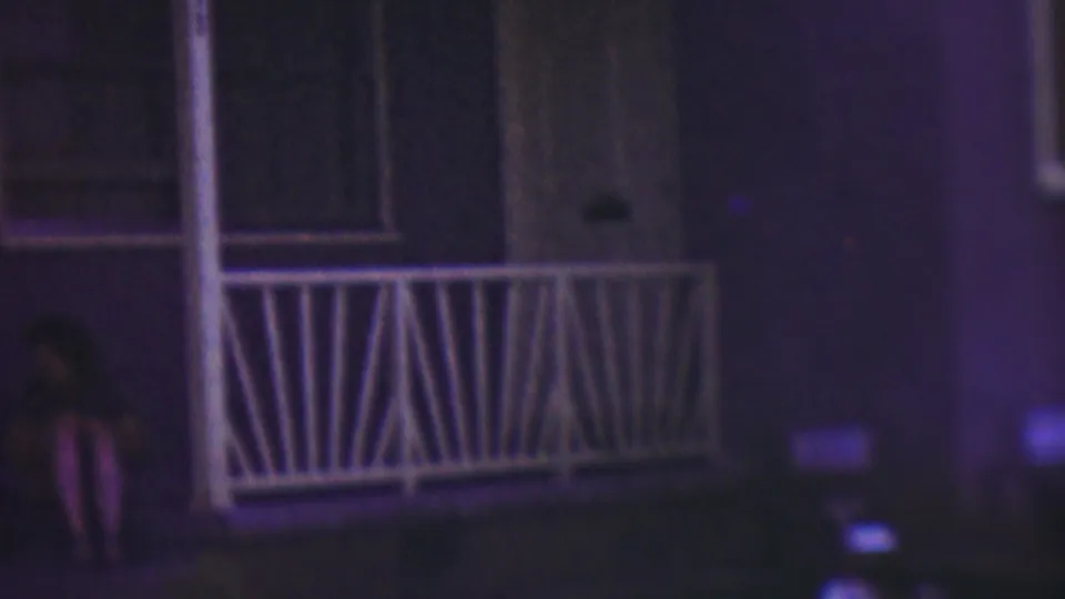 The woman is more centered on the front steps of the Cleveland house, seated beside the porch railing and doorway in soft, grainy 1953 8mm nighttime footage.