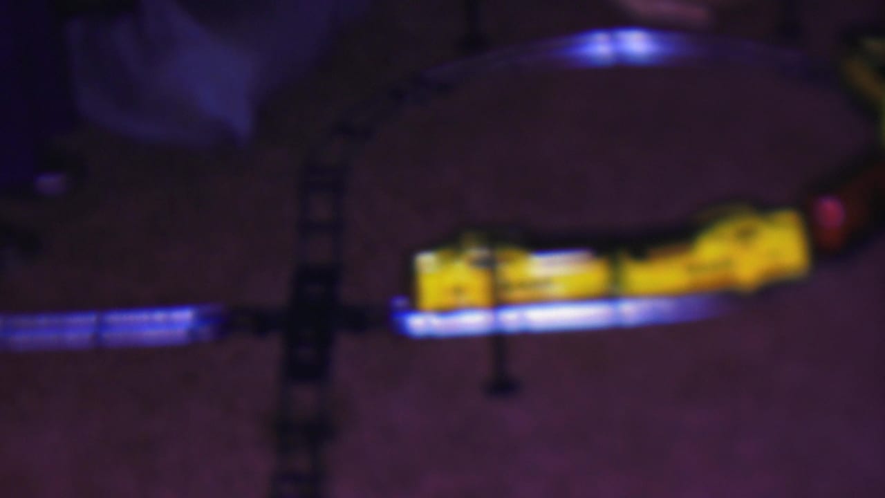 The yellow toy locomotive moves through the lower half of the 1953 Cleveland home-movie frame, with blue light glowing along the curved track on the carpet.