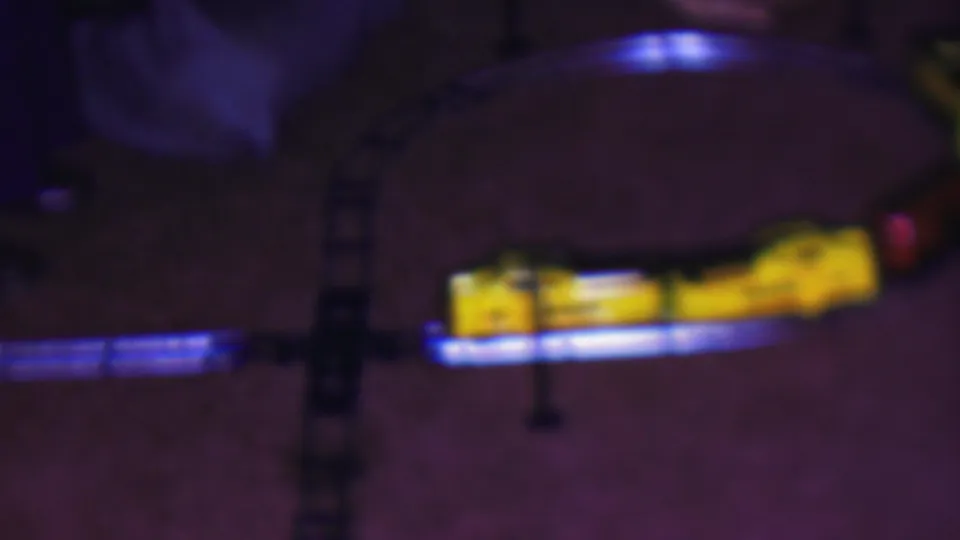 The yellow toy locomotive moves through the lower half of the 1953 Cleveland home-movie frame, with blue light glowing along the curved track on the carpet.