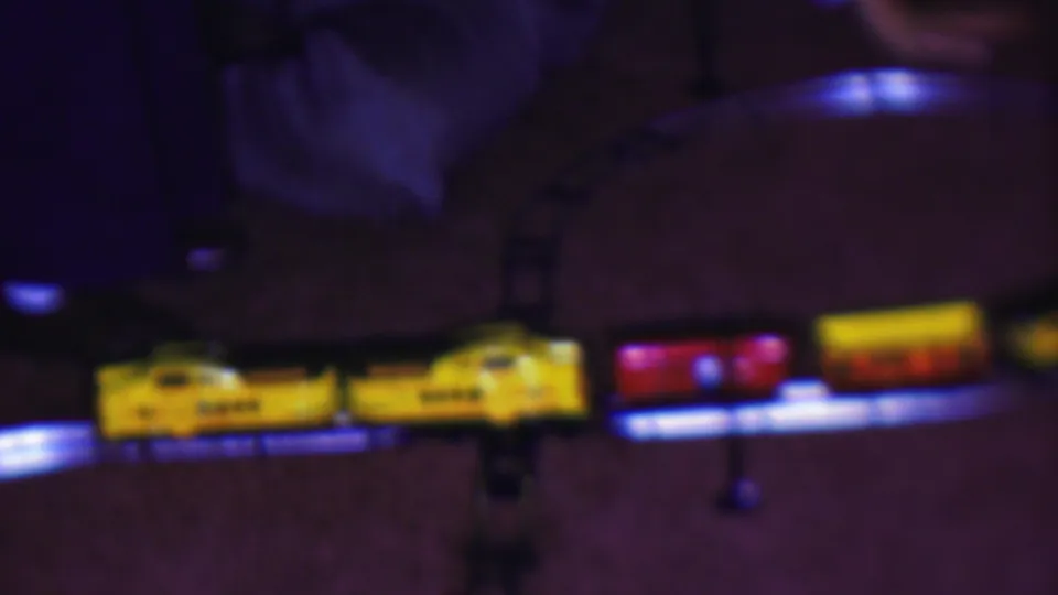 A yellow toy locomotive pulls red and yellow passenger cars along the curved track, captured in soft low-light 8mm texture inside a private interior.