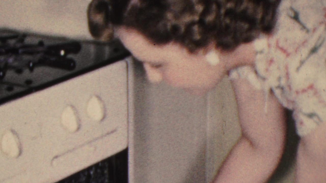 In a 1953 Cleveland home-movie frame, a woman in a floral dress bends beside a white stove while reaching toward the oven area in a private kitchen.