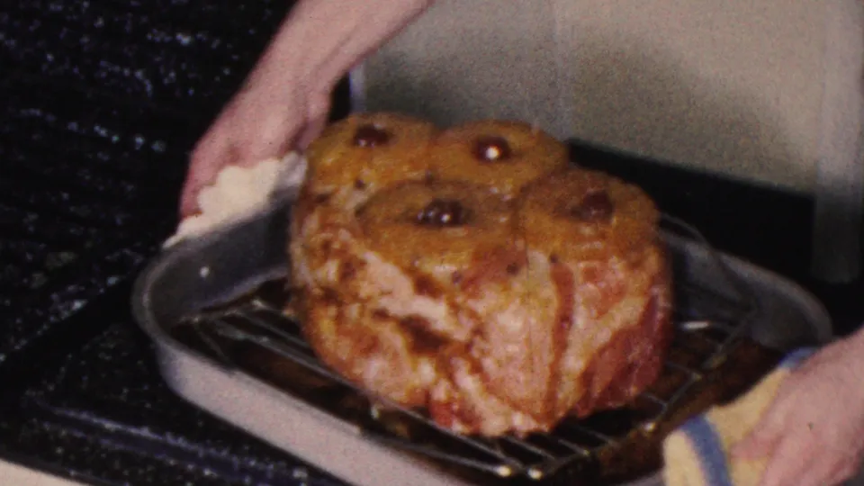 The 1953 home-movie frame shows a golden-brown glazed ham on a rack in a metal roasting pan, with a hand and potholder at the side of the stove.