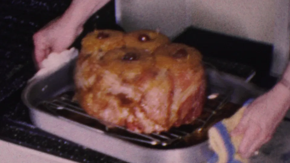 A woman in the Cleveland 1953 kitchen footage uses potholders to pull a metal roasting pan with a glazed ham from the stove oven.