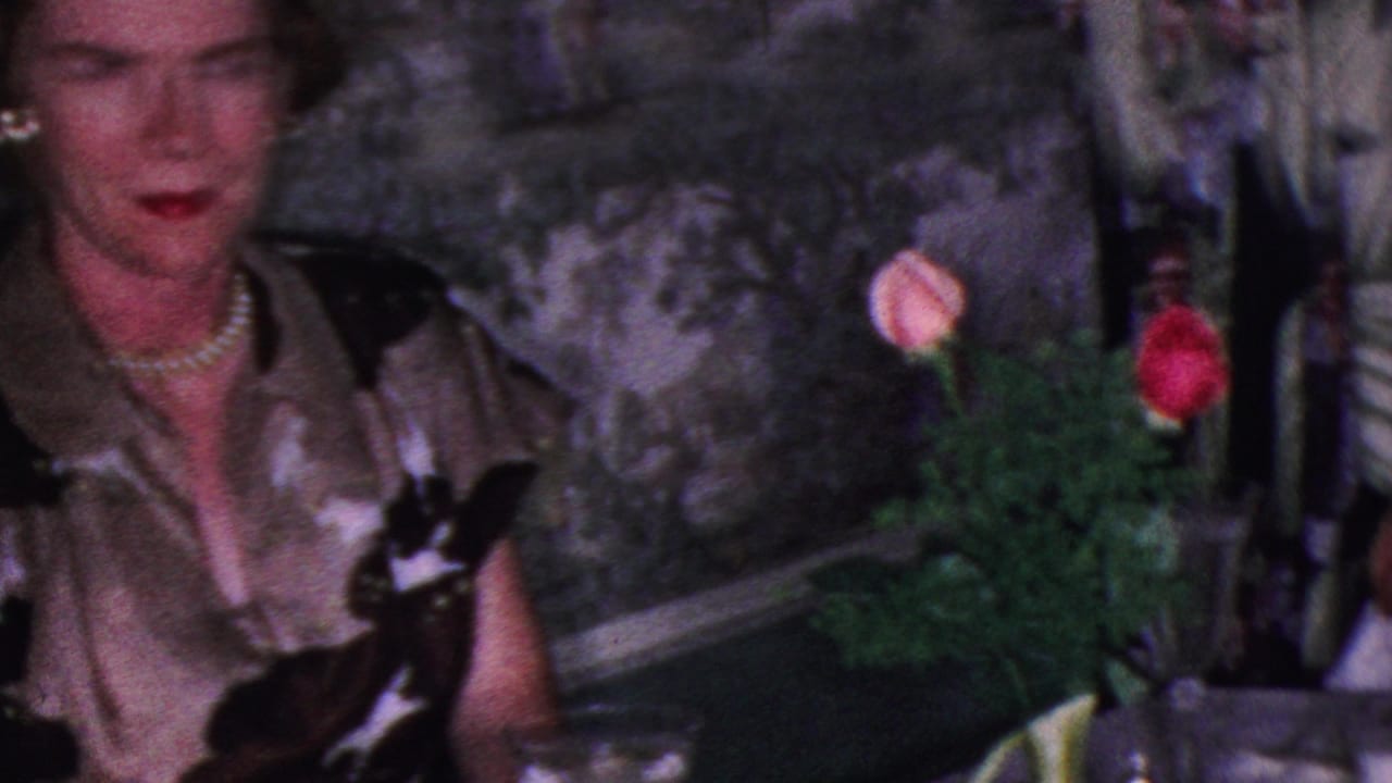 In this 1953 Cleveland home-movie frame, a woman in a patterned dress and pearl necklace sits at a formal dinner table with flowers, glassware, and food visible nearby.