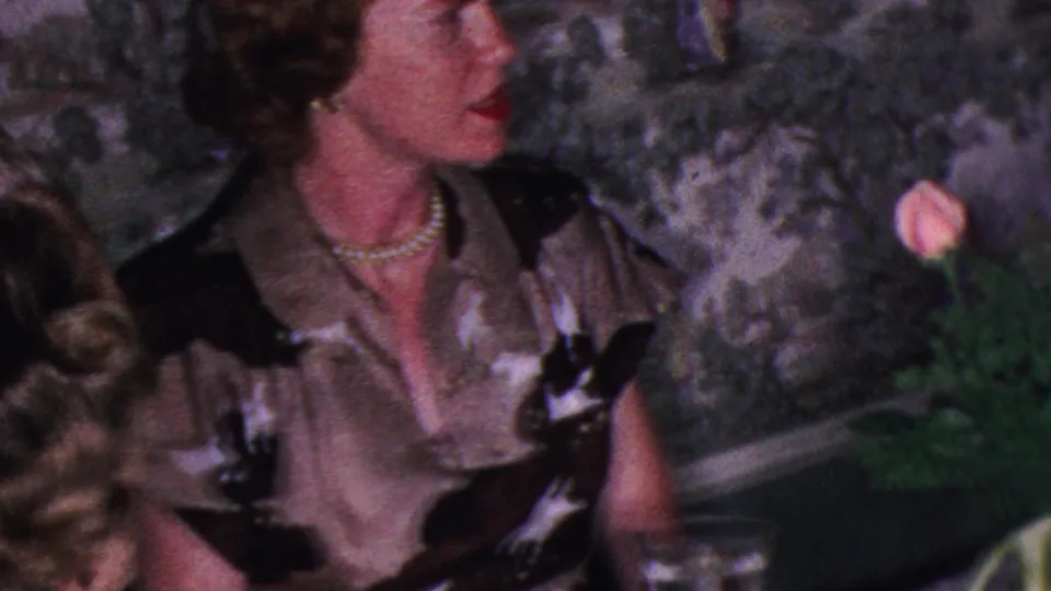 A woman in a patterned dress turns beside the dinner table in this Cleveland 1953 home-movie frame, with flowers, water glasses, food, and a partly visible shorter figure below the tabletop.