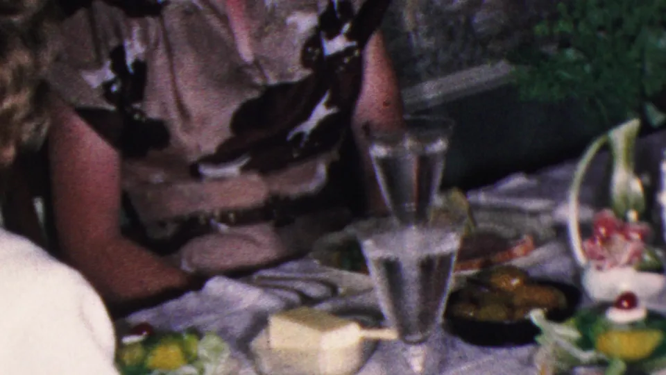 This 1953 Cleveland dinner-party frame shows a seated woman partly cropped beside a white tablecloth, water glass, plates, a pale rectangular food dish, and a small floral or vegetable garnish.