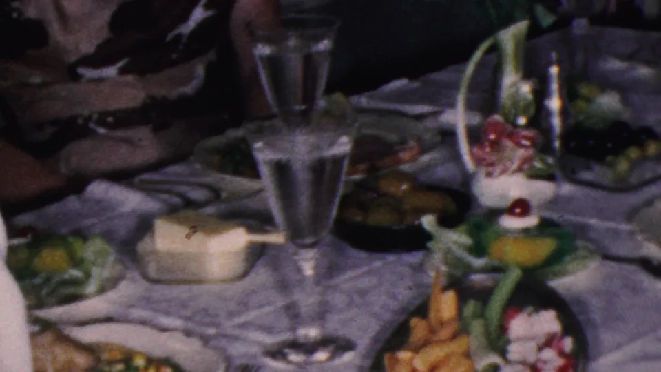 A formal dinner table in the 1953 Cleveland home-movie scene holds water glasses, plates of food, a pale dish, flowers, and decorative greenery, with a woman partly visible at the frame edge.