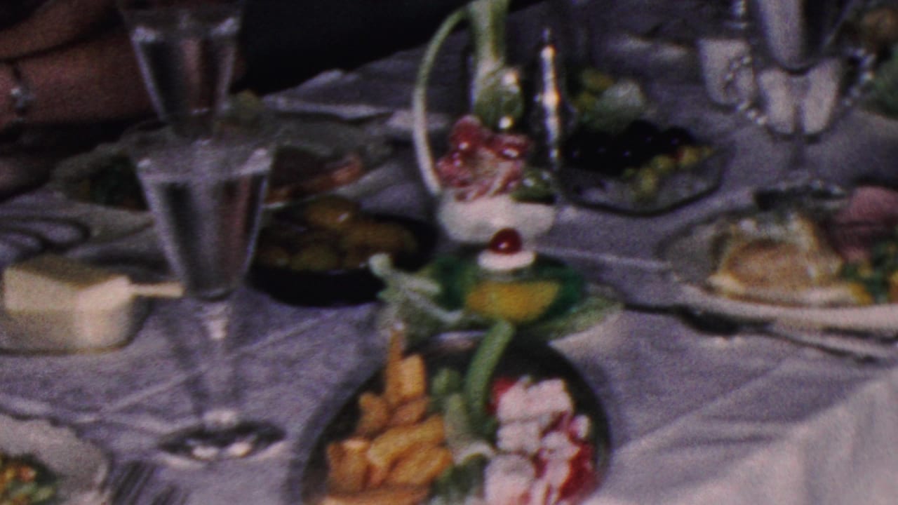 1953 Cleveland home-movie frame showing a formal dining table with a white tablecloth, goblets, serving dishes, olives, cutlery, and plated cold foods.