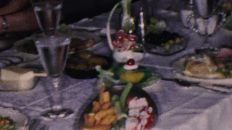 1953 Cleveland home-movie frame showing a formal dining table with a white tablecloth, goblets, serving dishes, olives, cutlery, and plated cold foods.