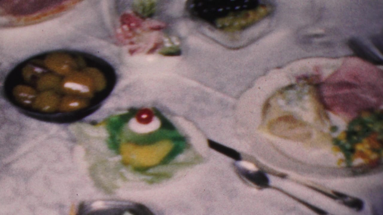 Dining table with plated ham, green dessert with cherry, olives, spoon, and white tablecloth.