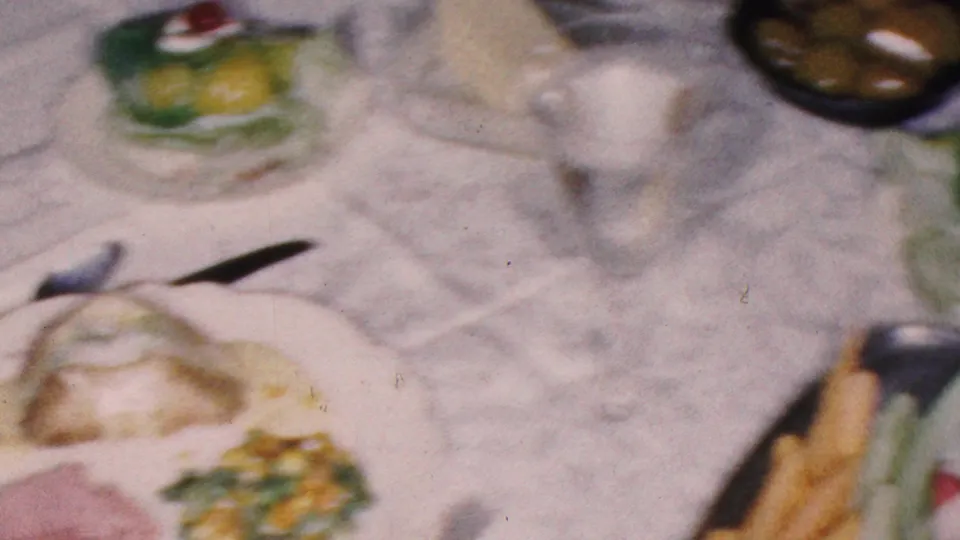 1953 Cleveland home-movie frame of a woman in profile seated near a formal dinner table with green desserts, serving dishes, cutlery, and a white tablecloth.