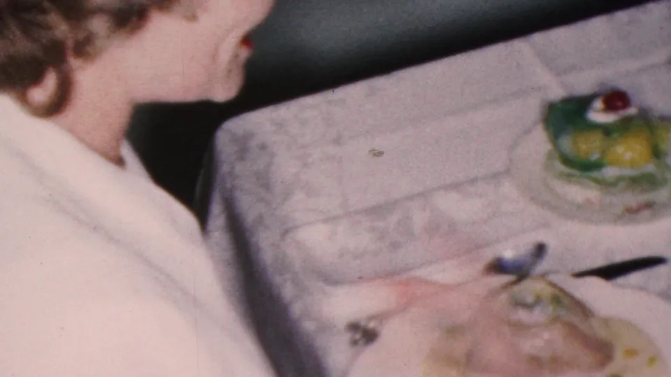 1953 Cleveland home-movie frame showing a woman in profile talking at a formal dining table with plated food, cutlery, goblets, and green desserts with cherry toppings.