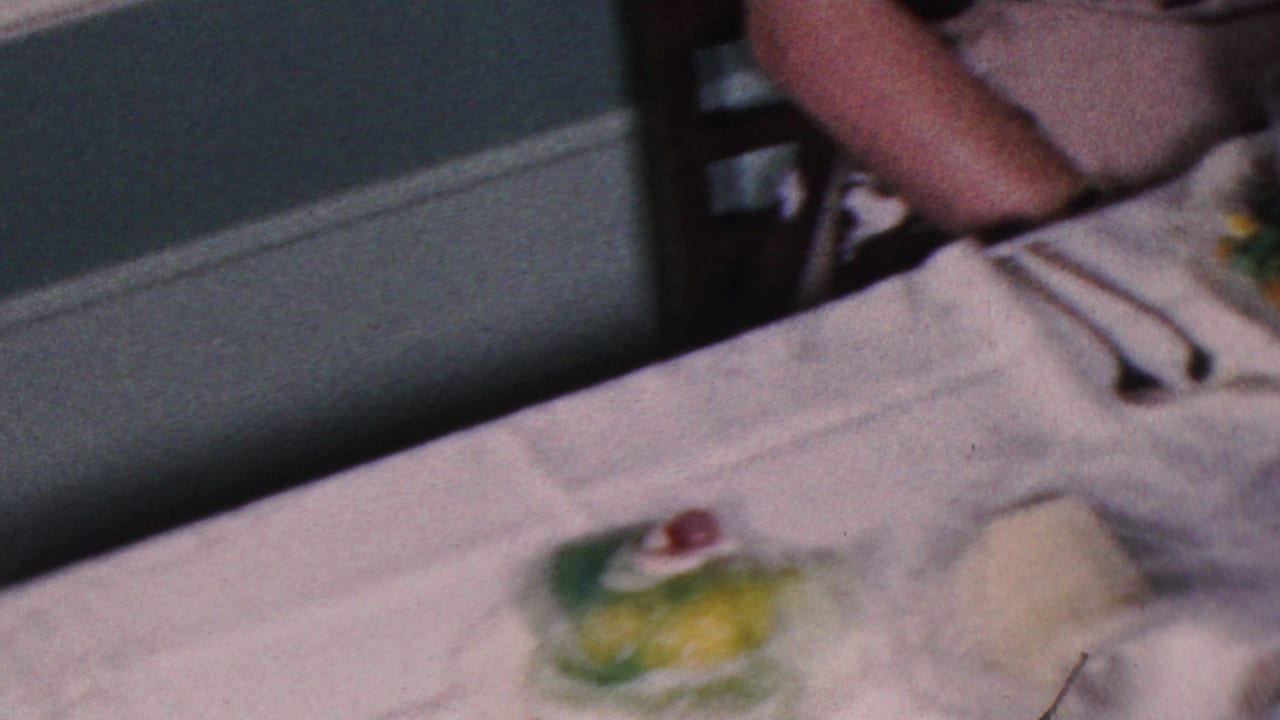 A formal dinner table in the Cleveland home-movie footage shows white linens, utensils, green gelatin desserts, and a partially visible diner seated at the edge of the frame.