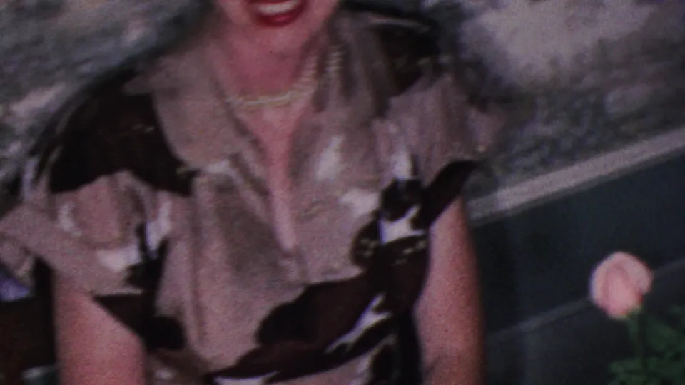 A woman wearing a patterned blouse and pearl necklace sits beside the dinner table in this 1953 Cleveland 8mm home-movie frame, with a flower and formal table setting nearby.