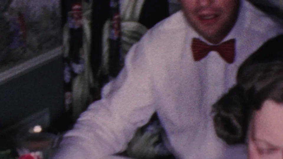 A man in a white shirt and red bow tie sits at the dinner table in the 1953 Cleveland home-movie scene, with other adult diners cropped around him.