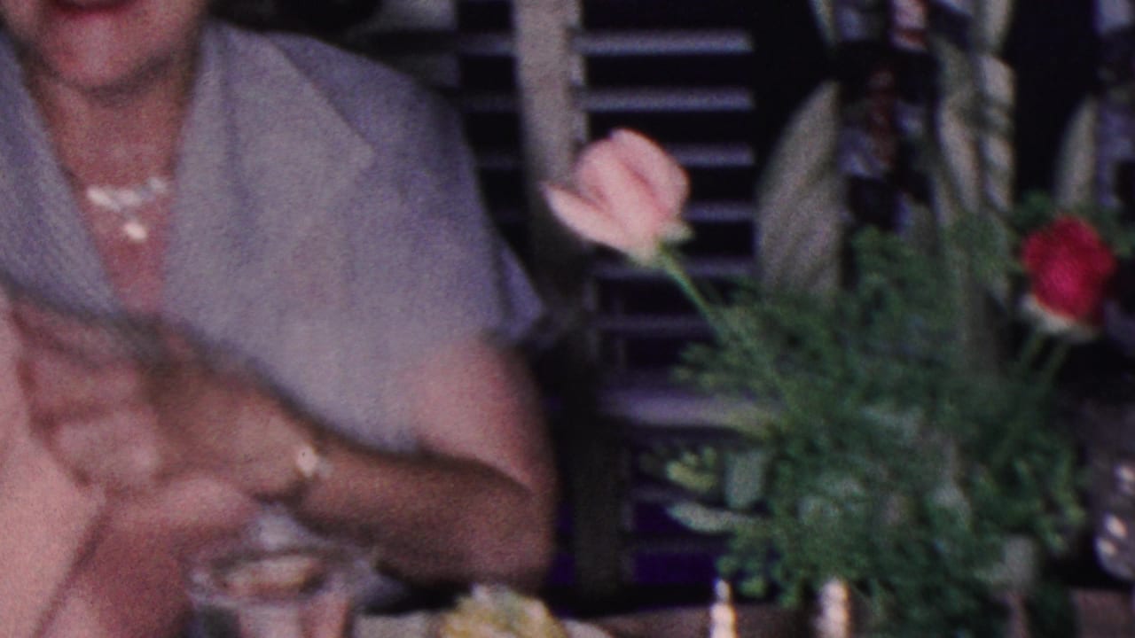 In this 1953 Cleveland home-movie frame, an adult woman in a light short-sleeve dress sits at a dinner table with flowers, glassware, and another partially visible guest nearby.