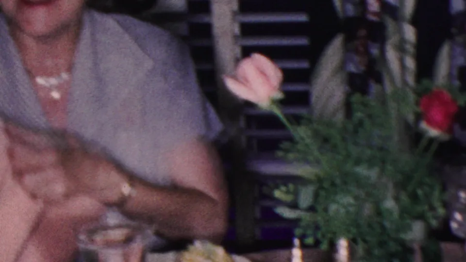 In this 1953 Cleveland home-movie frame, an adult woman in a light short-sleeve dress sits at a dinner table with flowers, glassware, and another partially visible guest nearby.