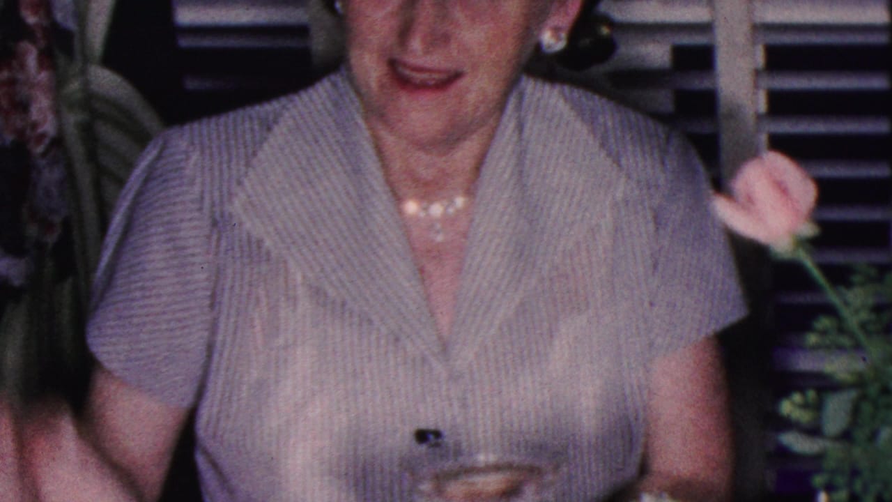 Smiling adult woman at a dinner table with flowers and dark window blinds behind her.