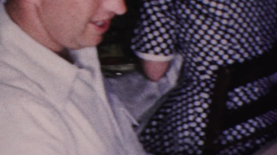 Two adult men in light shirts sit at a dinner gathering in the 1953 Cleveland footage, with a woman in a black-and-white polka dot dress partly visible beside them.