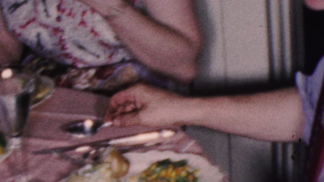 1953 Cleveland home-movie frame showing adults at a family dinner table, including a man in a striped shirt and red tie beside a floral-dress wearer, with plates and metal cups visible.