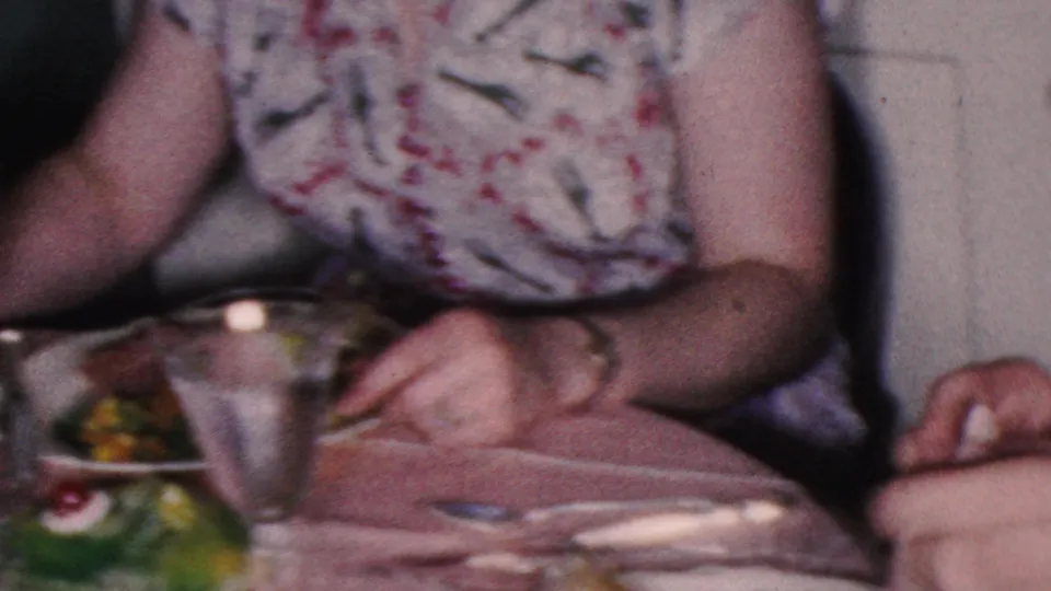 1953 Cleveland home-movie frame focusing on hands using cutlery at a dinner table with plates of food, a metal cup, and a patterned dress partially visible.