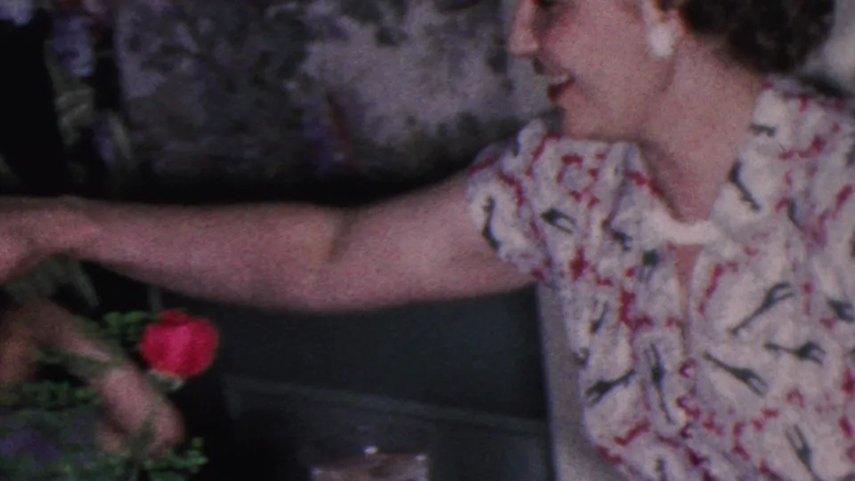 1953 Cleveland home-movie frame showing a woman in a floral dress reaching across the table near a red rose, with another person partly visible at left.