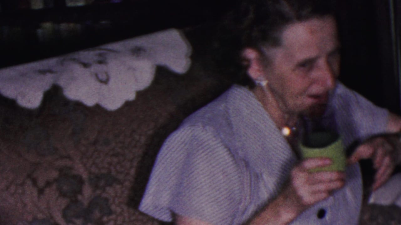 In a 1953 Cleveland home-movie frame, an older woman in a light striped blouse and pearl jewelry sits on a patterned couch while holding a green cup.
