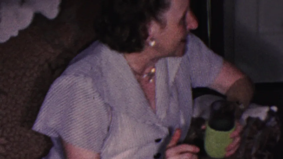 The woman turns in profile toward someone off camera while holding a green cup, seated against the patterned couch in the private interior.