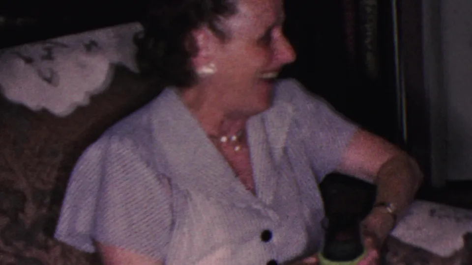 The woman smiles and looks off to the side from the couch, wearing pearl jewelry and a light blouse as the green cup rests near her hand.