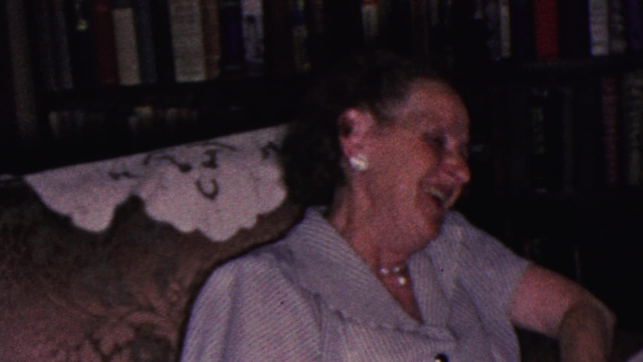 In a 1953 Cleveland home-movie frame, an elderly woman in a light blouse and pearl jewelry laughs on a patterned couch with bookshelves behind her.
