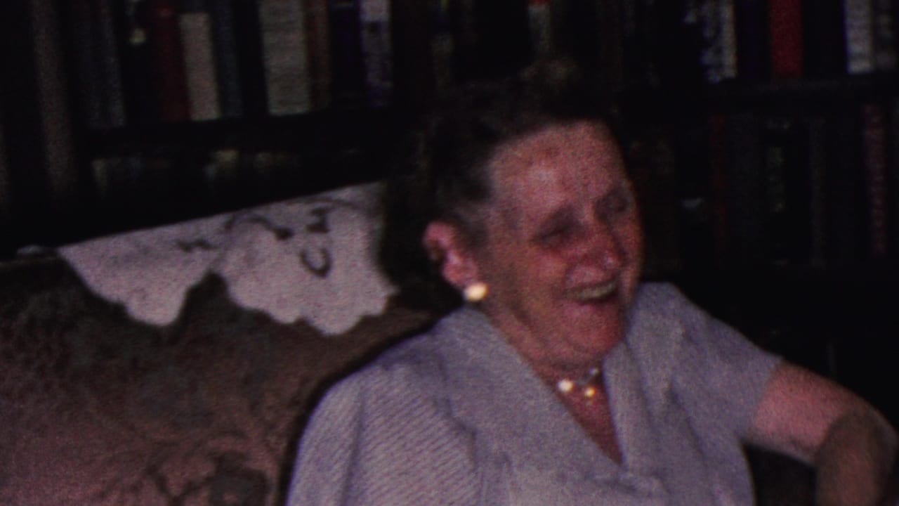 Woman smiling broadly on a couch with a white doily and bookshelves behind her.