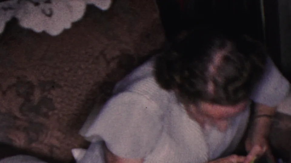 A second adult figure bends into the foreground of the 1953 home-movie frame, partly obscuring the couch while the domestic setting remains visible.