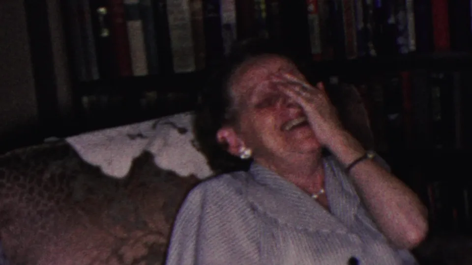 The woman laughs with her hand over her face in the Cleveland living room, framed by the couch, doily, and dark bookshelves.