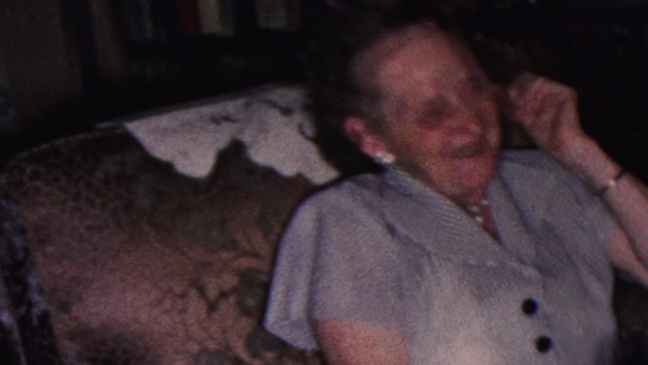 A woman in a striped dress and pearl jewelry laughs while seated on a patterned couch in a Cleveland home interior, consistent with the supplied 1953 8mm metadata.