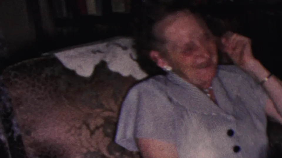 A woman in a striped dress and pearl jewelry laughs while seated on a patterned couch in a Cleveland home interior, consistent with the supplied 1953 8mm metadata.