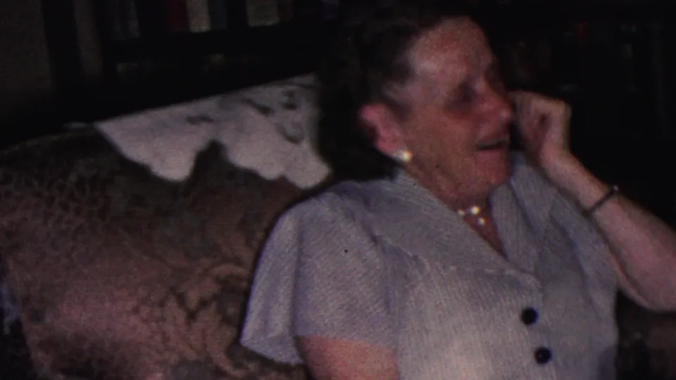The seated woman turns toward the side with her hand raised near her face, surrounded by patterned upholstery and dark shelving in the Cleveland home-movie interior.